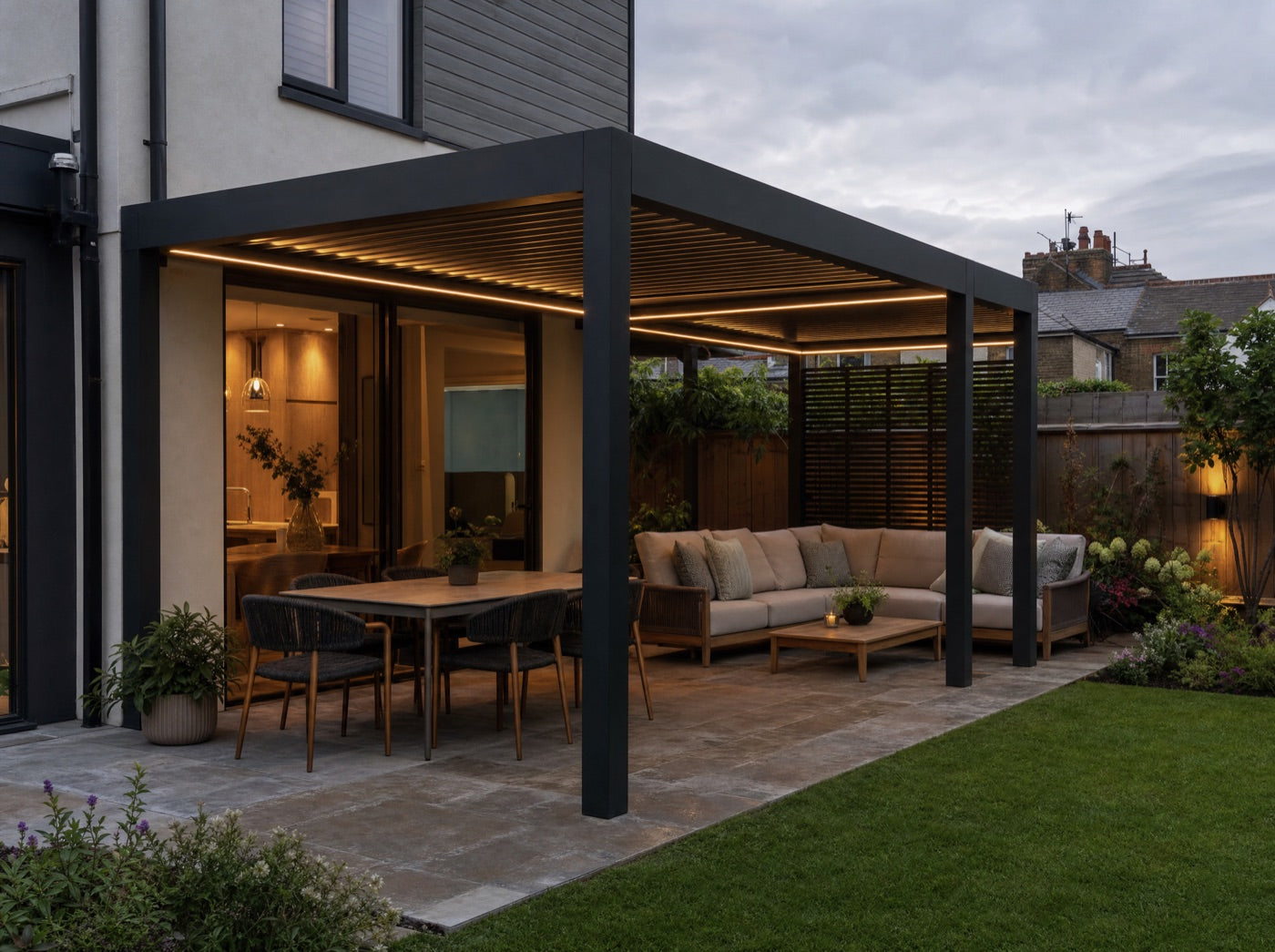 An outdoor sofa and dining set beneath a louvred aluminium pergola in a garden at dusk